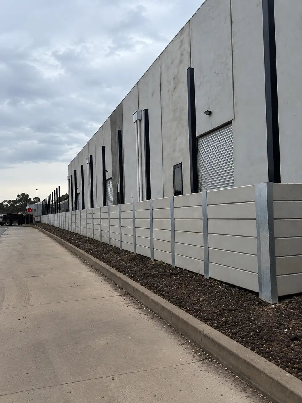 Grey concrete sleeper retaining wall on galvanised steel posts along a driveway beside a warehouse