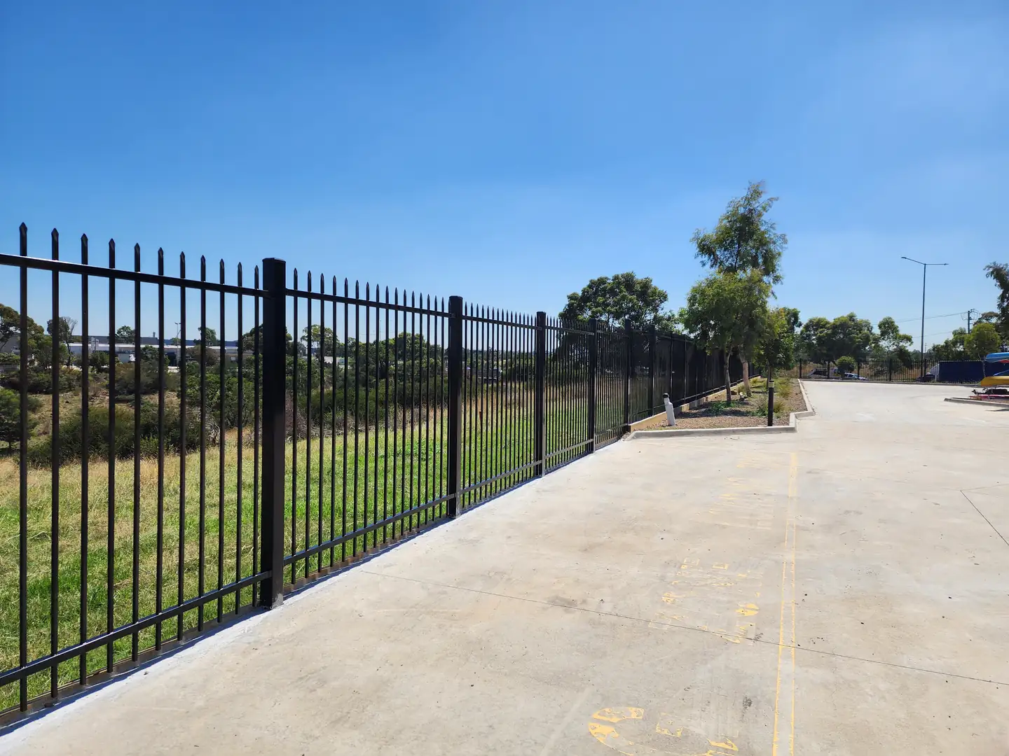 New black steel spear-top fence running along the edge of a wide concrete yard, with long grass on the other side